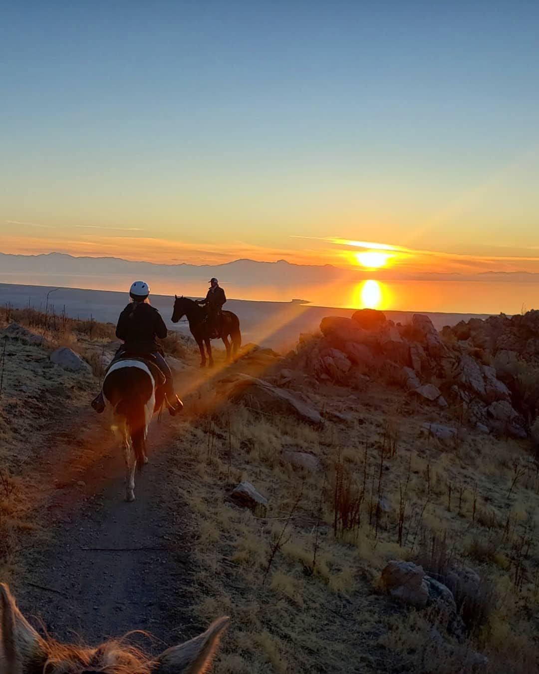 Two people riding horses on a trail at sunrise with mountains in the distance.