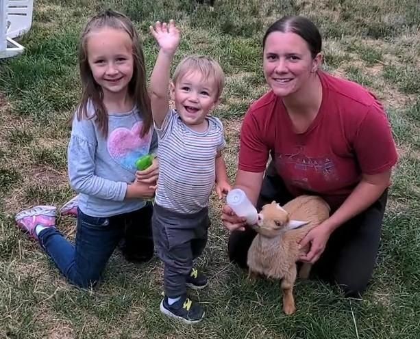 Woman and two children outdoors feeding a small goat with a bottle on grass.